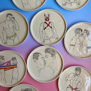 Może przedstawiać: Kolekcja dziewięciu okrągłych talerzy ceramicznych z czarnymi ilustracjami liniowymi przedstawiającymi postacie w różnych pozach, w tym osoby w czerwonych maskach i uprzężach, całujące się postacie oraz postacie w tęczowej bieliźnie.