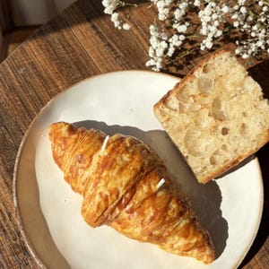 May include: A flaky, golden-brown croissant candle and a piece of bread are on a white plate. The croissant has two white wicks. The plate is on a wooden surface, with white flowers in the background.