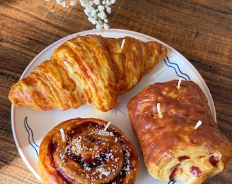 May include: A plate with three baked goods: a croissant, a pastry roll, and a cinnamon roll. Each has a candle wick. The plate has a pink rim with a blue wave design. The background is a wooden table with white flowers.