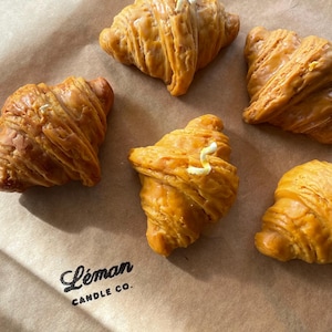 May include: Five golden-brown croissant-shaped candles with a butter-like topping are arranged on a brown paper surface. The candles are from Leman Candle Co. and are designed to resemble baked goods.
