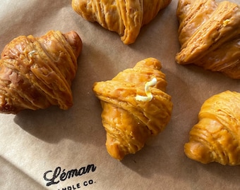 May include: Five golden-brown croissant-shaped candles with a butter-like topping are arranged on a brown paper surface. The candles are from Leman Candle Co. and are designed to resemble baked goods.