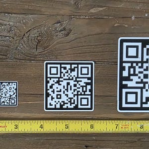 May include: Four black and white QR code stickers of varying sizes are displayed on a wooden surface. A yellow measuring tape is placed below the stickers, showing measurements in inches. The largest sticker is approximately 4 inches square.