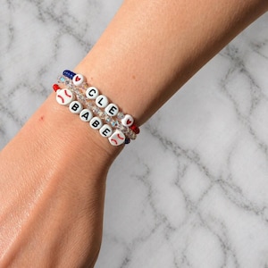 May include: Three beaded bracelets with white letter beads spelling "CLE BABE" and baseball-themed accents. The bracelets feature red, white, and blue beads, with baseball and heart charms. The bracelets are worn on a wrist.