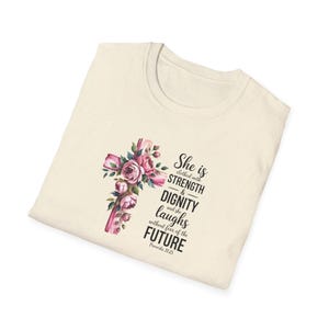 May include: A cream-coloured t-shirt with a floral cross design in pink and green. The shirt has the text "She is clothed with STRENGTH & DIGNITY and she laughs without fear of the FUTURE" from Proverbs 31:25.