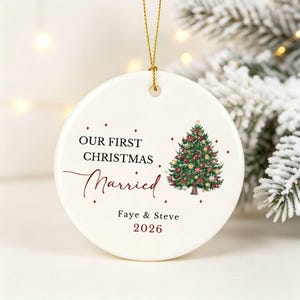 Può includere: Un ornamento rotondo in ceramica bianca appeso a un cordino dorato. Presenta un'illustrazione di un albero di Natale verde con palline rosse e dorate, e il testo "OUR FIRST CHRISTMAS Married", "Faye & Steve" e "2026" in rosso scuro.
