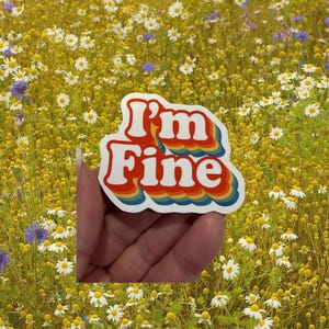 May include: A white sticker with the text "I'm Fine" in red, outlined in a rainbow gradient. The sticker is held in front of a field of yellow flowers with white and purple accents.