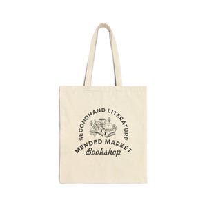 May include: A natural-colored canvas tote bag featuring a black graphic print. The design includes a stack of books and floral elements surrounded by the text "SECONDHAND LITERATURE MENDED MARKET Bookshop" arranged in a circular and linear layout.
