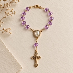 Op de afbeelding: Een goudkleurige rozenkransarmband met gefacetteerde paarse kralen, een centrale ovale bedel met een wit midden omringd door kleine heldere steentjes en een goudkleurige crucifixhanger aan een fijne ketting.