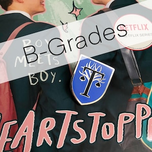 May include: A blue enamel pin with a silver border and a white letter "T" in the centre. The letter "T" is surrounded by a stylised tree with a silver trunk and leaves. The pin is on a book cover with the text "Boy Meets Boy" and "Heartstopper" on it.