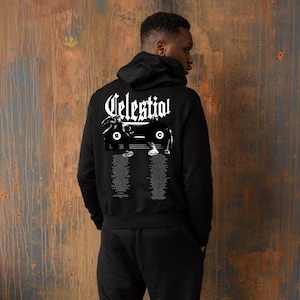 Puede incluir: Sudadera con capucha negra con la palabra "Celestial" en letras góticas blancas en la capucha. La parte trasera de la sudadera con capucha presenta un diseño gráfico con texto adicional debajo. La persona lleva un pantalón de chándal negro.