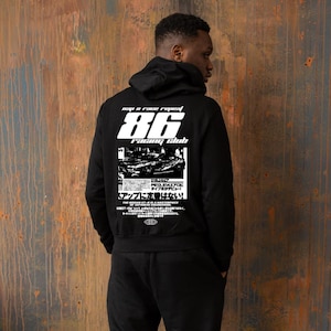 Puede incluir: Sudadera con capucha negra con texto gráfico blanco y una imagen de coche en la espalda. El texto dice "86 Racing Club" y "Pap a race repeat". La sudadera la lleva una persona delante de una pared de metal oxidado.