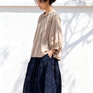 May include: A woman wearing a beige linen blouse with a tie-neck detail and three-quarter sleeves. She is wearing a long, navy blue plaid skirt. The outfit is completed with woven sandals. The background features a white wall and a mountain range.