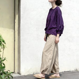 May include: A person wearing a purple long-sleeved top and beige trousers. The top has a round neckline and gathered sleeves. The trousers are loose-fitting with a drawstring waist. The person is wearing straw sandals.