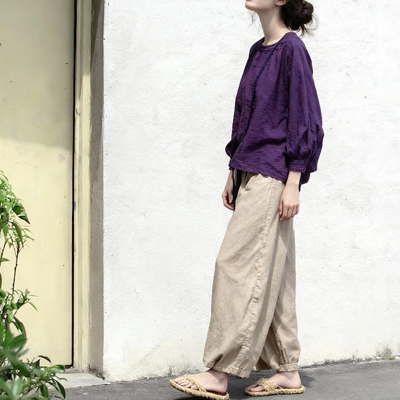 May include: A person wearing a purple long-sleeved top and beige trousers. The top has a round neckline and gathered sleeves. The trousers are loose-fitting with a drawstring waist. The person is wearing straw sandals.