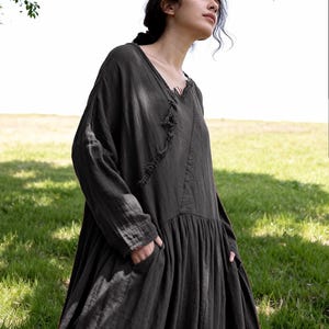 May include: A long, dark grey dress with long sleeves and a V-neck. The dress has a loose, flowing silhouette with a gathered skirt and pockets. The fabric appears to be a lightweight, textured material, suitable for warmer weather.