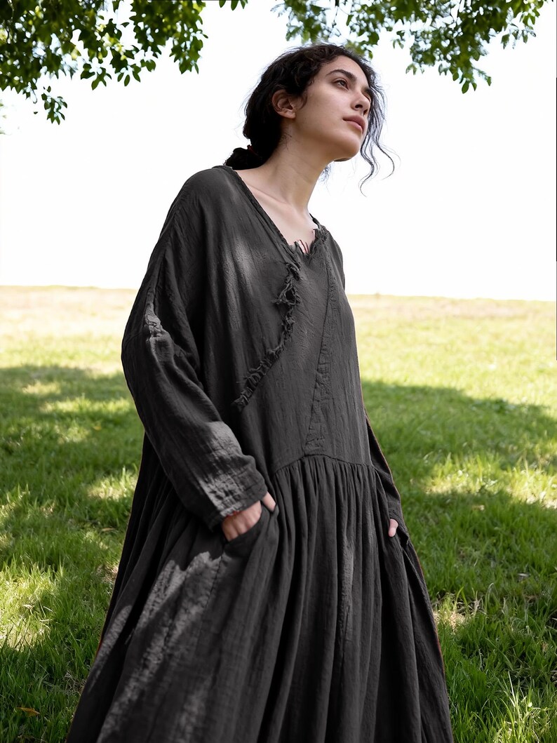 May include: A long, dark grey dress with long sleeves and a V-neck. The dress has a loose, flowing silhouette with a gathered skirt and pockets. The fabric appears to be a lightweight, textured material, suitable for warmer weather.