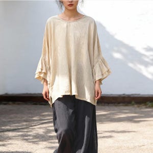 May include: A beige long-sleeved blouse featuring a round neckline and tiered, ruffled bell sleeves. The garment has a loose, flowing fit and is paired with dark charcoal gray wide-leg trousers that have a visible textured fabric finish.