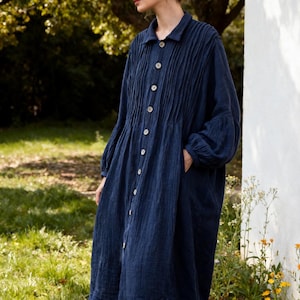 May include: A long-sleeved, dark blue button-down dress featuring a collared neckline and vertical pleats extending from the shoulders to the waist. The garment has a loose, flowing silhouette with gathered cuffs and visible side pockets.