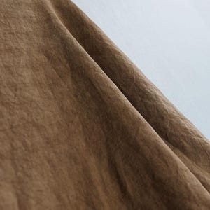 May include: Close-up of a brown fabric with a textured surface. The fabric is folded, creating shadows and highlights that emphasize its texture. The background is a light, neutral color.