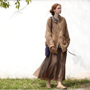 May include: A woman wearing a brown linen jacket with embroidered details, a long brown skirt, and woven sandals. She is also wearing a green shirt and carrying a purple bag. The outfit is set against a white wall and green grass.