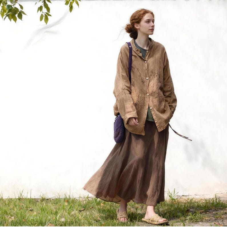 May include: A woman wearing a brown linen jacket with embroidered details, a long brown skirt, and woven sandals. She is also wearing a green shirt and carrying a purple bag. The outfit is set against a white wall and green grass.