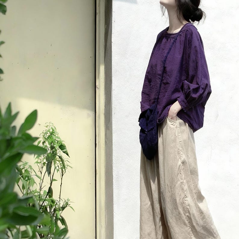 May include: A casual outfit comprising a long-sleeved, purple blouse and beige wide-leg trousers. The blouse has a round neckline and gathered sleeves. The trousers are a light, neutral colour. The outfit is styled in a relaxed manner.