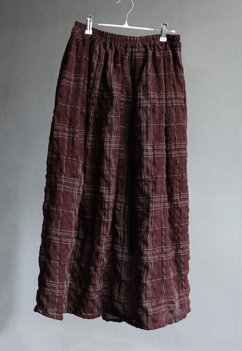 May include: A long, burgundy skirt with a white grid pattern, hanging from a hanger. The skirt has an elastic waistband and a flowing silhouette, suitable for casual wear.