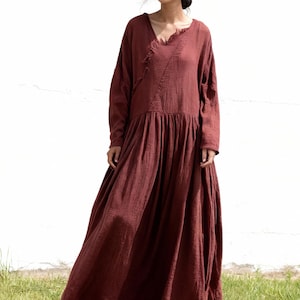 May include: A full-length maroon dress with long sleeves and a V-neck. The dress features a gathered waist and a flowing skirt. The model is standing on green grass against a white wall. The dress is made of a soft, textured material.