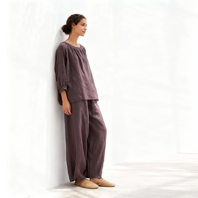 May include: A two-piece set of loose-fitting, dusty purple linen clothing. The top has a round neckline and three-quarter sleeves. The trousers are wide-legged. The outfit is paired with woven, tan-coloured slippers. The model is leaning against a white wall.