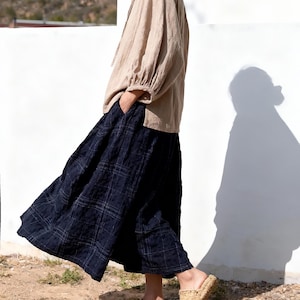 May include: A beige linen blouse with long sleeves and a tie-neck detail is paired with a navy blue plaid skirt. Woven straw slip-on sandals complete the outfit. The ensemble is set against a white wall and a natural outdoor setting.