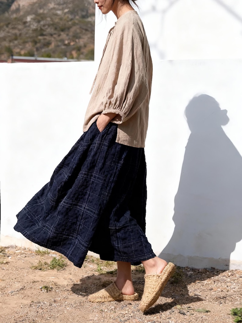 May include: A beige linen blouse with long sleeves and a tie-neck detail is paired with a navy blue plaid skirt. Woven straw slip-on sandals complete the outfit. The ensemble is set against a white wall and a natural outdoor setting.