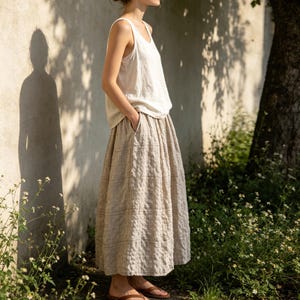 May include: A woman wearing a cream-colored tank top and a beige, textured midi skirt with pockets. The outfit is paired with brown sandals. The clothing appears to be made of linen or a similar natural fabric, suitable for warm weather.