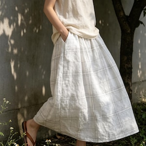 May include: A white plaid skirt with a wide, flowing design. The skirt has a high waist and pockets. The person in the image is wearing a cream-colored top and brown sandals. The skirt's plaid pattern is made of thin, dark gray lines.