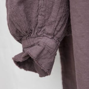 May include: Close-up of a mauve-coloured garment sleeve. The sleeve has a gathered, puffed design at the shoulder and a ruffled cuff. The fabric appears to be a textured linen or similar material, with visible seams and folds.