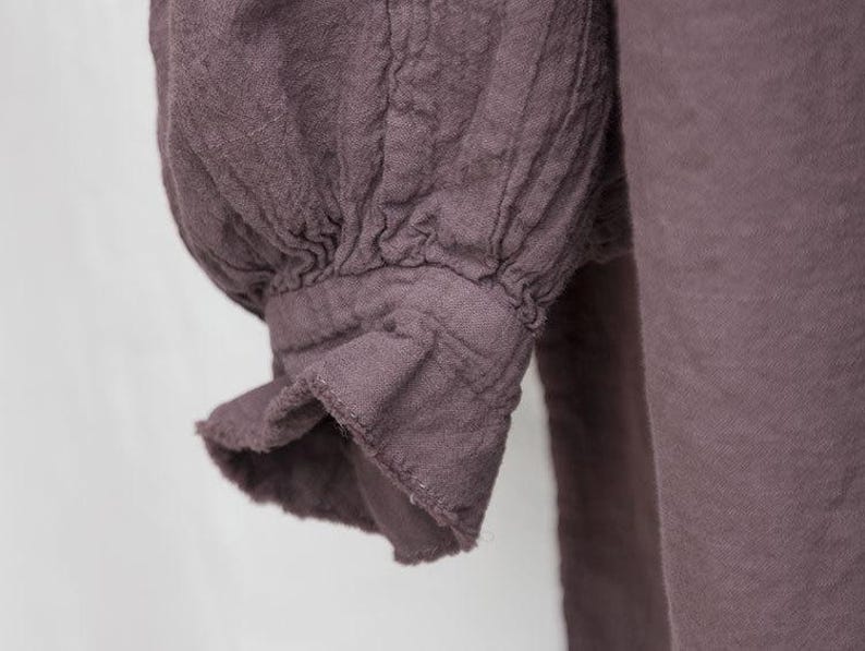 May include: Close-up of a mauve-coloured garment sleeve. The sleeve has a gathered, puffed design at the shoulder and a ruffled cuff. The fabric appears to be a textured linen or similar material, with visible seams and folds.