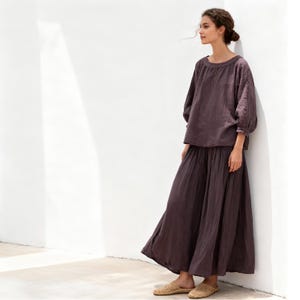 May include: A two-piece outfit in a muted purple hue. The ensemble includes a long, flowing skirt and a top with a round neckline and three-quarter sleeves. The outfit is paired with woven sandals. The model is leaning against a white wall.