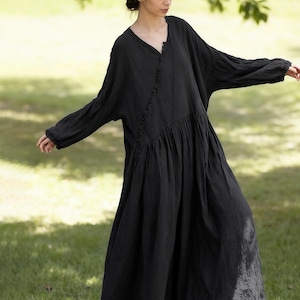 May include: A full-length black dress with a V-neck and long sleeves. The dress features a gathered waist and a ruffled trim down the front. The model is standing on grass, arms extended, in a natural setting.