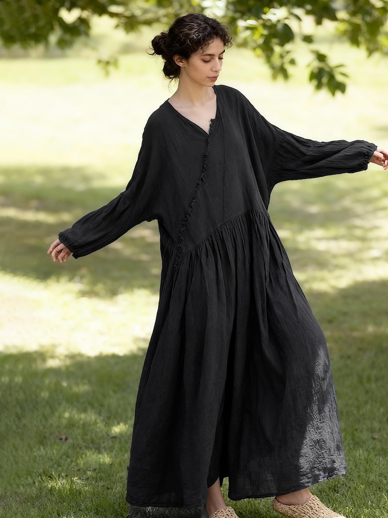 May include: A full-length black dress with a V-neck and long sleeves. The dress features a gathered waist and a ruffled trim down the front. The model is standing on grass, arms extended, in a natural setting.