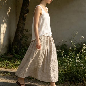 May include: A woman wearing a white tank top and a beige plaid skirt. The skirt is a midi length and has a pleated design. The outfit is made of a lightweight fabric, suitable for warm weather. The woman is walking outside near a tree and some flowers.