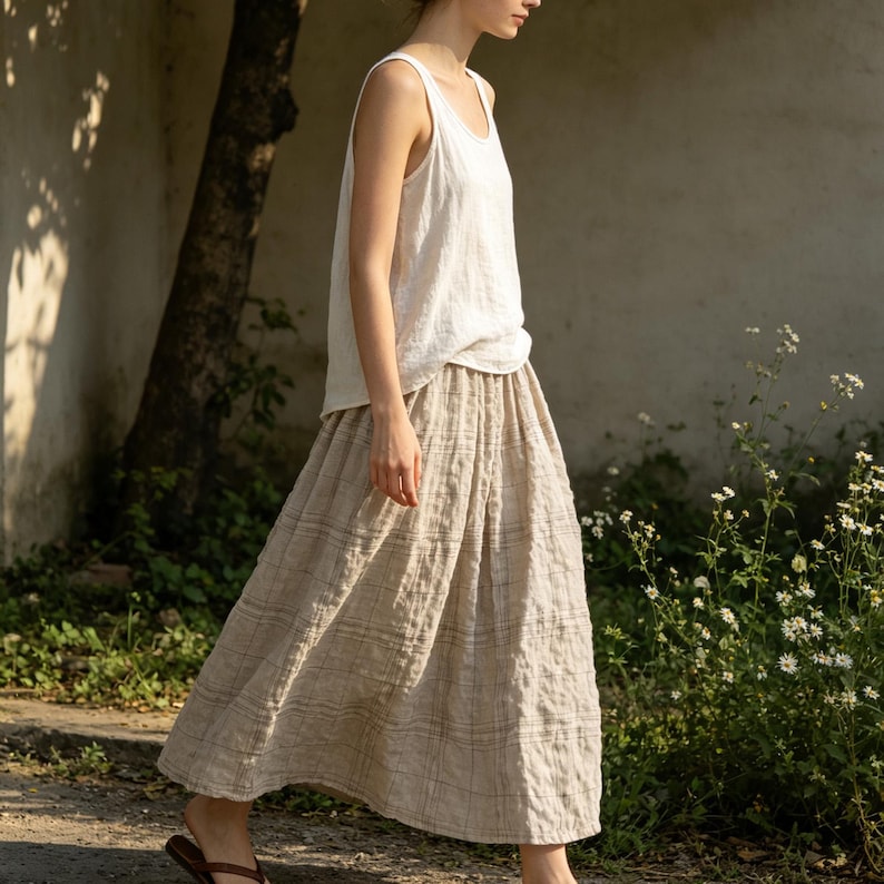 May include: A woman wearing a white tank top and a beige plaid skirt. The skirt is a midi length and has a pleated design. The outfit is made of a lightweight fabric, suitable for warm weather. The woman is walking outside near a tree and some flowers.
