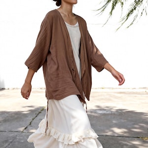 May include: A brown linen cardigan with three-quarter sleeves is worn over a cream-colored dress with a ruffled hem. The cardigan is open-fronted and drapes loosely. The dress has a simple, elegant design, suitable for casual wear.