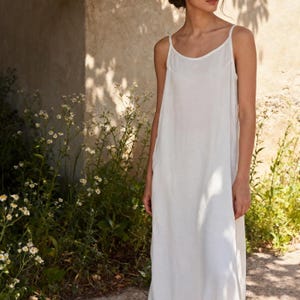 Könnte beinhalten: Ein langes, weißes Slip-Kleid mit Spaghettiträgern. Das Kleid hat ein schlichtes, fließendes Design. Das Model steht vor einer Steinmauer mit Grün und Sonnenlicht im Hintergrund. Das Kleid scheint aus einem leichten Stoff zu sein.