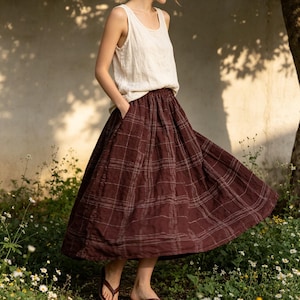 May include: A woman wearing a cream-colored tank top and a burgundy plaid skirt. The skirt has a flowing, A-line silhouette with a subtle grid pattern. The outfit is casual and comfortable, suitable for warm weather. The skirt has pockets.