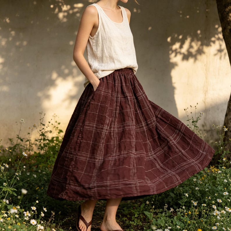 May include: A woman wearing a cream-colored tank top and a burgundy plaid skirt. The skirt has a flowing, A-line silhouette with a subtle grid pattern. The outfit is casual and comfortable, suitable for warm weather. The skirt has pockets.