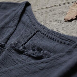 May include: Close-up of a dark grey, textured garment. The fabric appears to be a soft, woven material with visible stitching and frayed edges. The neckline is visible, and a dried leaf rests in the background.