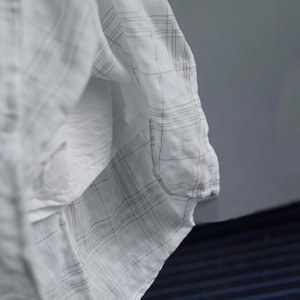 May include: Close-up of a white fabric with a subtle plaid pattern in black. The fabric is draped, creating soft folds and shadows. The material appears lightweight and possibly linen or cotton.