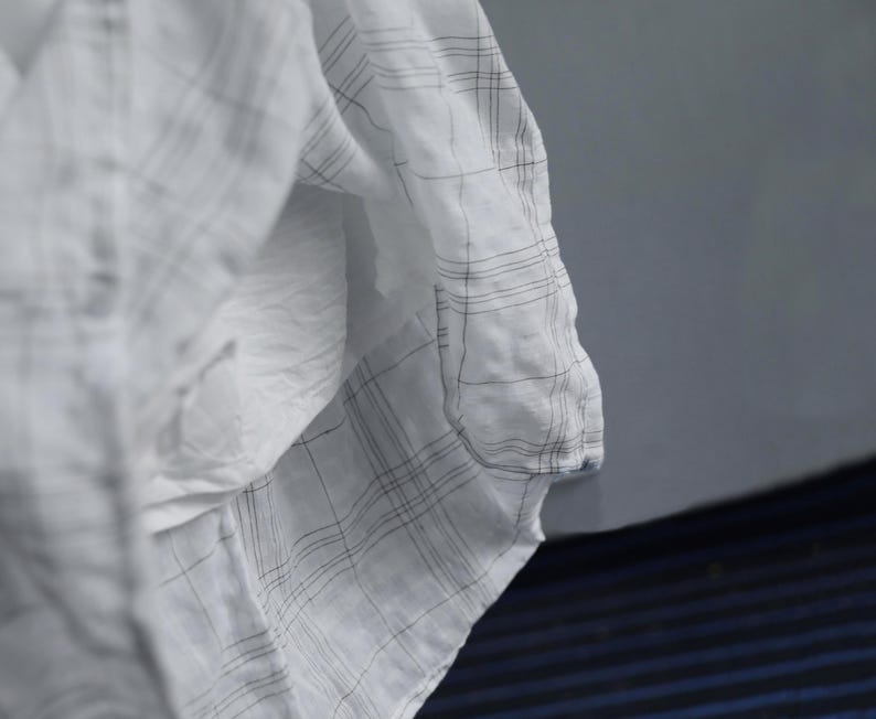 May include: Close-up of a white fabric with a subtle plaid pattern in black. The fabric is draped, creating soft folds and shadows. The material appears lightweight and possibly linen or cotton.