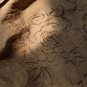 May include: Close-up of a tan fabric with a floral pattern. The design features a large rose and other botanical elements in a darker brown color. The fabric is slightly wrinkled, with shadows adding depth.