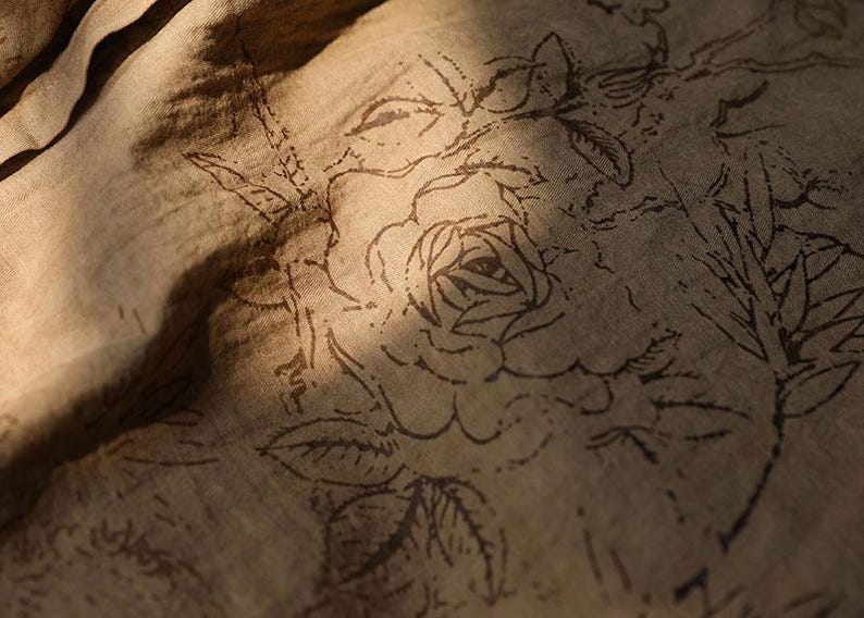 May include: Close-up of a tan fabric with a floral pattern. The design features a large rose and other botanical elements in a darker brown color. The fabric is slightly wrinkled, with shadows adding depth.