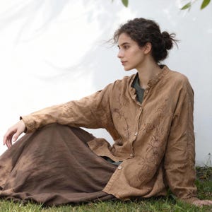 May include: A woman wearing a tan linen shirt with floral embroidery and a long brown skirt, sitting on the grass. The shirt is buttoned and has a mandarin collar. The outfit is casual and earthy.